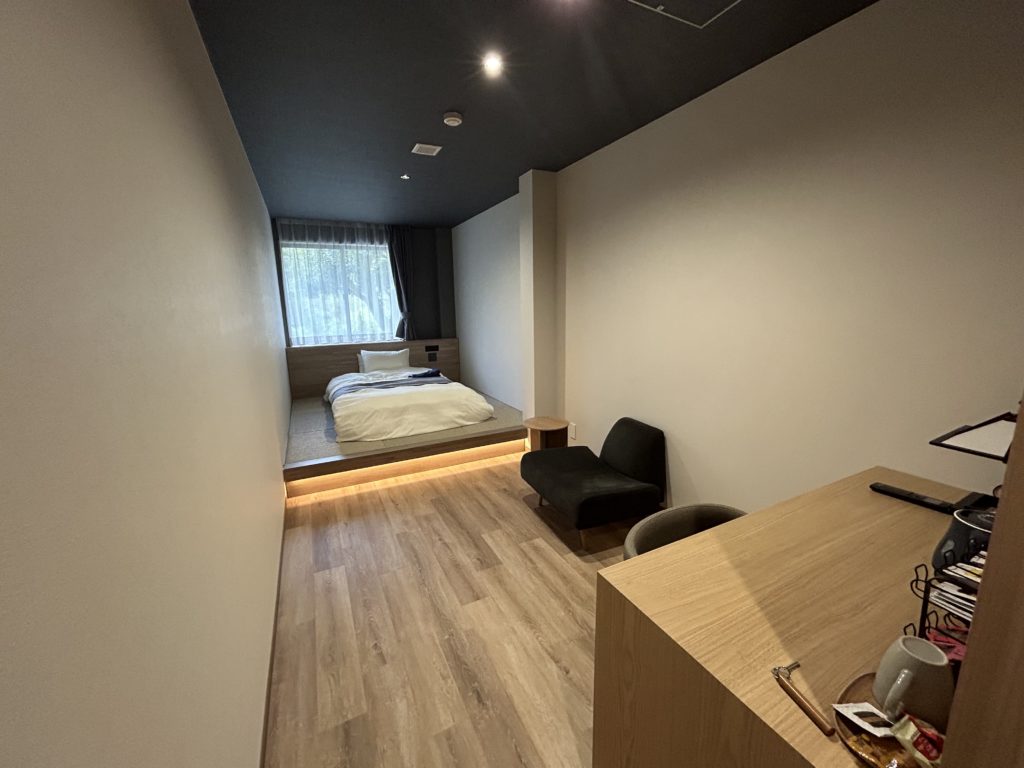 室内の様子。洋室で、手前に机、真ん中に一人掛けのソファ、奥にベッドが配置されている。